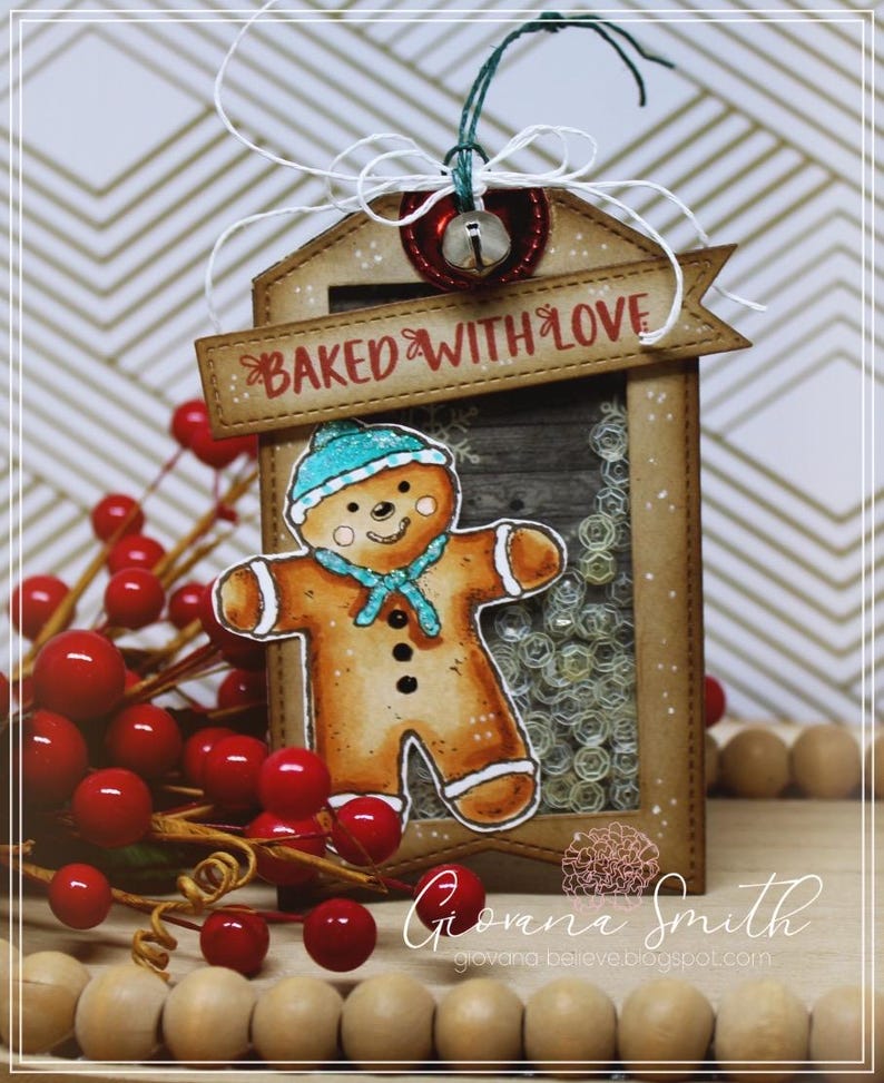 May include: A handmade Christmas tag featuring a gingerbread man with a blue hat and scarf. The tag reads "BAKED WITH LOVE" and includes a small bell and red berries. The tag is brown with a clear window.