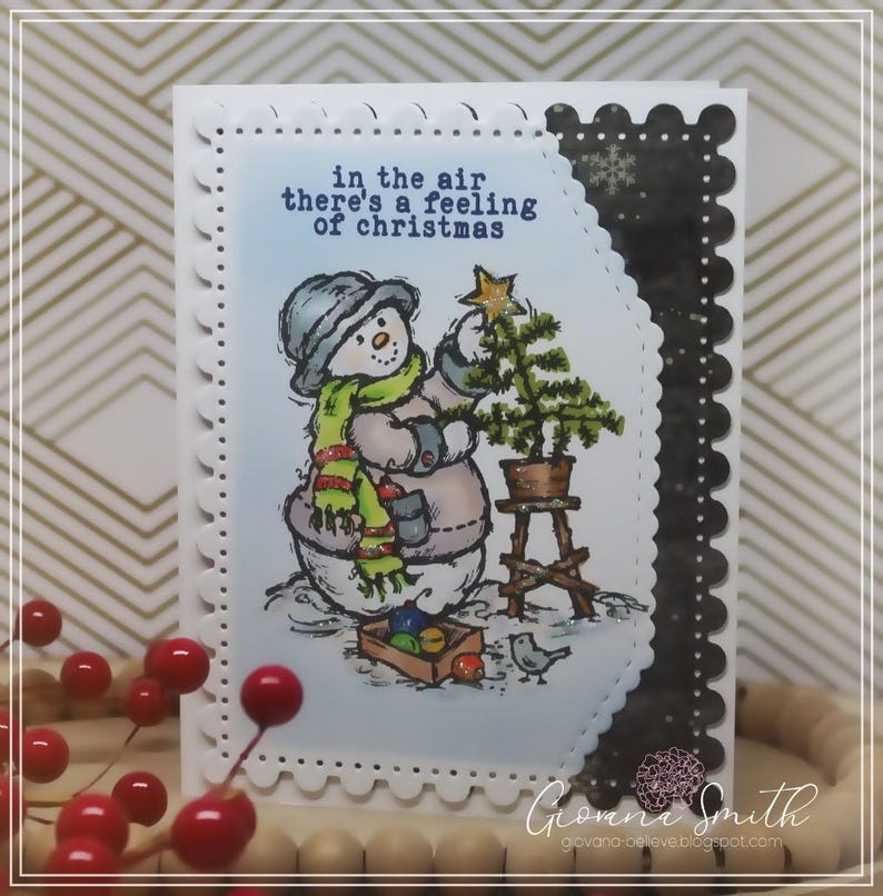 May include: A Christmas-themed greeting card featuring a snowman decorating a small Christmas tree. The snowman wears a hat, scarf, and mittens. The card has the text "in the air there's a feeling of christmas". The card has a scalloped edge.