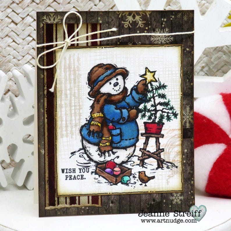 May include: A Christmas card featuring a snowman in a blue coat and brown hat, placing a star on a small Christmas tree. The card has a wooden frame and the text "WISH YOU PEACE."
