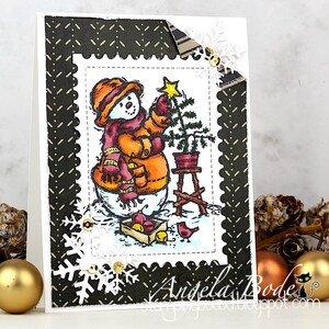 May include: A handmade Christmas card featuring a snowman decorating a small Christmas tree. The snowman wears an orange hat, scarf, and mittens. The card has a black and white patterned border with snowflake and sequin embellishments. The card is decorated with a small bird and a crate of apples.