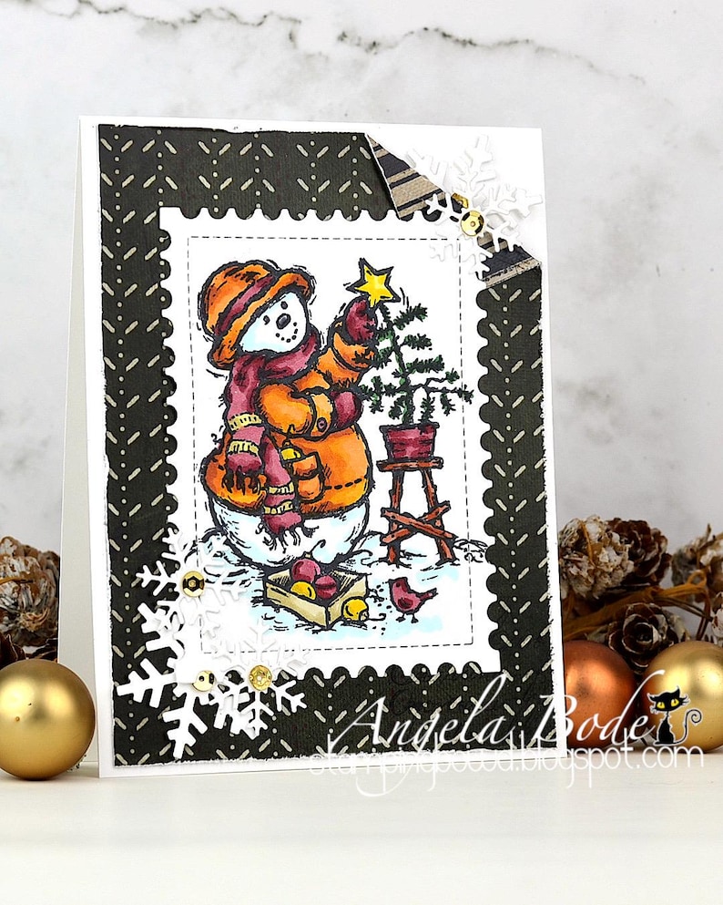 May include: A handmade Christmas card featuring a snowman decorating a small Christmas tree. The snowman wears an orange hat, scarf, and mittens. The card has a black and white patterned border with snowflake and sequin embellishments. The card is decorated with a small bird and a crate of apples.