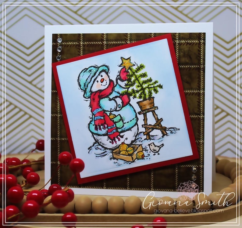 May include: A handmade Christmas card featuring a snowman holding a star and decorating a small tree. The snowman wears a blue hat, red scarf, and mittens. The card has a plaid background and a red border.