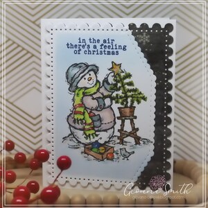 May include: A handmade Christmas card featuring a snowman decorating a small Christmas tree. The snowman wears a hat, scarf, and mittens. The card has the text "in the air there's a feeling of christmas". The card has a scalloped edge and a black decorative border.