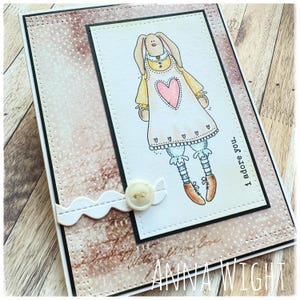 May include: A greeting card with a cartoon bunny wearing a dress with a heart on it. The card has a brown background with white polka dots and the text "I adore you".