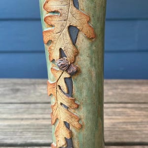 May include: A ceramic vase with a green glaze and a design of brown leaves and acorns. The vase is handmade and has a unique, organic shape.