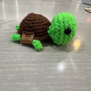 Peut inclure: Une tortue crochet&eacute;e &agrave; la main, avec une carapace marron et une t&ecirc;te, des pieds et une queue verts. La tortue a un &oelig;il en bouton noir et une petite &eacute;tiquette avec l'inscription "by Sarah L."