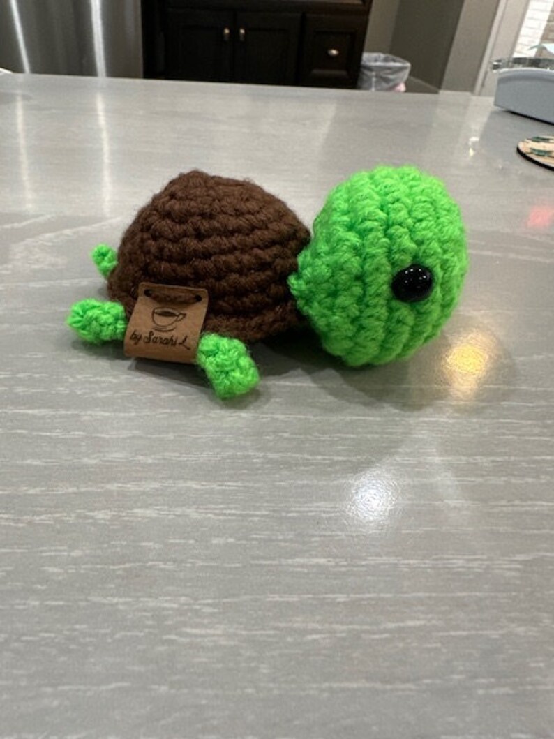 Peut inclure: Une tortue crochet&eacute;e &agrave; la main, avec une carapace marron et une t&ecirc;te, des pieds et une queue verts. La tortue a un &oelig;il en bouton noir et une petite &eacute;tiquette avec l'inscription "by Sarah L."