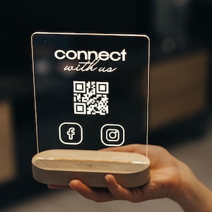 Puede incluir: Un letrero de acrílico transparente con el texto "connect with us" y un código QR. El letrero también tiene iconos para Facebook e Instagram. El letrero está sobre una base de madera.