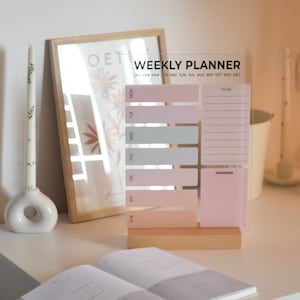 Puede incluir: Un planificador semanal de acrílico transparente con una base de madera. El planificador tiene un esquema de color rosa y gris e incluye secciones para cada día de la semana, listas de tareas y recordatorios. El texto en el planificador dice "Weekly Planner" y "Jan Feb Mar Apr May Jun Jul Aug Sep Oct Nov Dec".