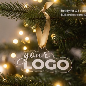 Pode incluir: Um ornamento de acrílico transparente com formato de nuvem apresentando o texto branco your LOGO, suspenso por uma fita de cetim dourada. O ornamento está pendurado num ramo de pinheiro verde decorado com luzes brancas quentes.