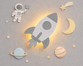 May include: A wall-mounted rocket ship light fixture in gray and white, emitting a warm glow. The set includes decorative wooden wall cutouts of an astronaut holding a flag, a crescent moon, a ringed planet, a striped planet, and several stars.