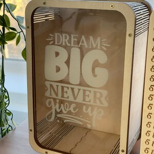 以下が含まれることがあります： 透明な前面パネルが付いたスーツケース型の木製貯金箱。前面パネルには「DREAM BIG NEVER give up」という文字が印刷されています。