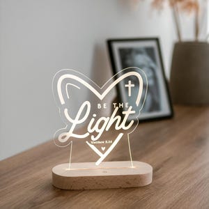 Könnte beinhalten: Ein weißes, herzförmiges Nachtlicht aus Acryl mit den Worten "Be the Light" und "Matthew 5:14" in weißer, kursiv geschriebener Schrift. Das Licht steht auf einem Holzsockel mit einem Schalter an der Seite.