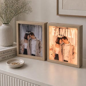 May include: Two wooden framed photo displays, one with a photo and the other with the word "FAMILY" illuminated. The photos show a couple. The frames are on a white surface with a vase of flowers and a small dish.