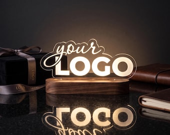 Puede incluir: Un letrero de acrílico iluminado personalizado con el texto "your LOGO" en luz blanca, montado sobre una base de madera oscura de forma ovalada y lisa. El letrero emite un brillo suave, creando un reflejo claro en la superficie oscura y pulida.