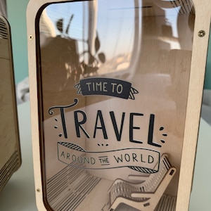 以下が含まれることがあります： 透明な前面パネルが付いた木製の旅行鞄。鞄には「Time to Travel Around the World」という黒と白のグラフィックが描かれています。鞄の中には、ミニチュアの木製椅子が入っています。