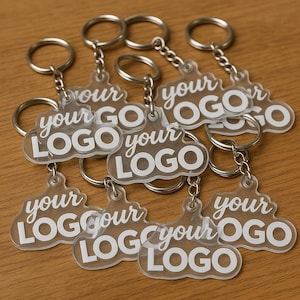 Puede incluir: Llaveros de acrílico transparente con anillas plateadas. Cada llavero tiene la inscripción "your LOGO" en blanco. Los llaveros están colocados sobre una superficie de madera marrón claro.