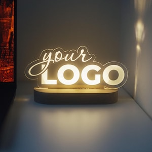 Pode incluir: Um letreiro de acrílico iluminado personalizado com o texto "your LOGO" em uma combinação de fontes cursivas e negrito sem serifa. O painel de acrílico transparente é montado em uma base de madeira escura que emite um brilho quente.