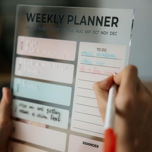 Könnte beinhalten: Ein klarer Wochenplaner aus Acryl mit den Worten "WEEKLY PLANNER" oben. Der Planer hat Abschnitte für jeden Tag und eine "TO DO"-Liste. Eine Hand schreibt mit einem rot-weißen Stift auf die "TO DO"-Liste.