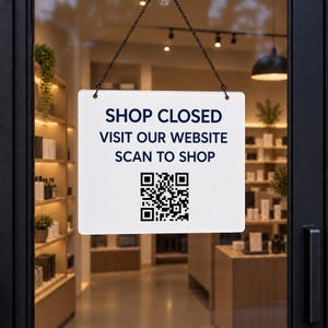 Könnte beinhalten: Ein rechteckiges weißes Schild, das an einer schwarzen Kette hängt, mit dunkelblauem Text, der SHOP CLOSED VISIT OUR WEBSITE SCAN TO SHOP lautet, und einem schwarz-weißen QR-Code unter dem Text, angebracht an einer Glastür.