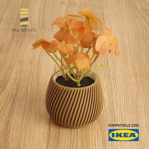 Può includere: Un vaso rotondo di colore marrone, con una texture, con una piccola pianta arancione al suo interno. Il vaso ha un design a coste. Il testo "COMPATIBLE CON: IKEA" è visibile nell'angolo in basso a destra dell'immagine.