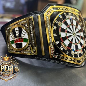 May include: A black and gold championship belt with a dartboard design. The belt features a central dartboard, surrounded by gold accents and the words "UNIVERSAL DARTS CHAMPION". The belt also has a logo that says "PEG CHAMPIONSHIP".