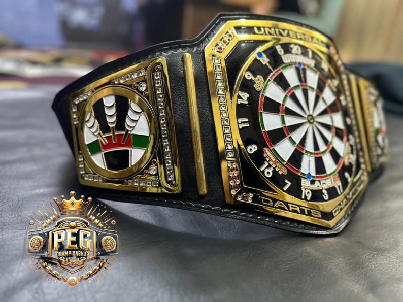 May include: A black and gold championship belt with a dartboard design. The belt features a central dartboard, surrounded by gold accents and the words "UNIVERSAL DARTS CHAMPION". The belt also has a logo that says "PEG CHAMPIONSHIP".