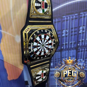 May include: A black and gold championship belt featuring a dartboard design at the center. The belt has gold accents, decorative emblems, and the text "UNIVERSAL WINMAU". The image also includes the text "PEG CHAMPIONSHIP BELTS".
