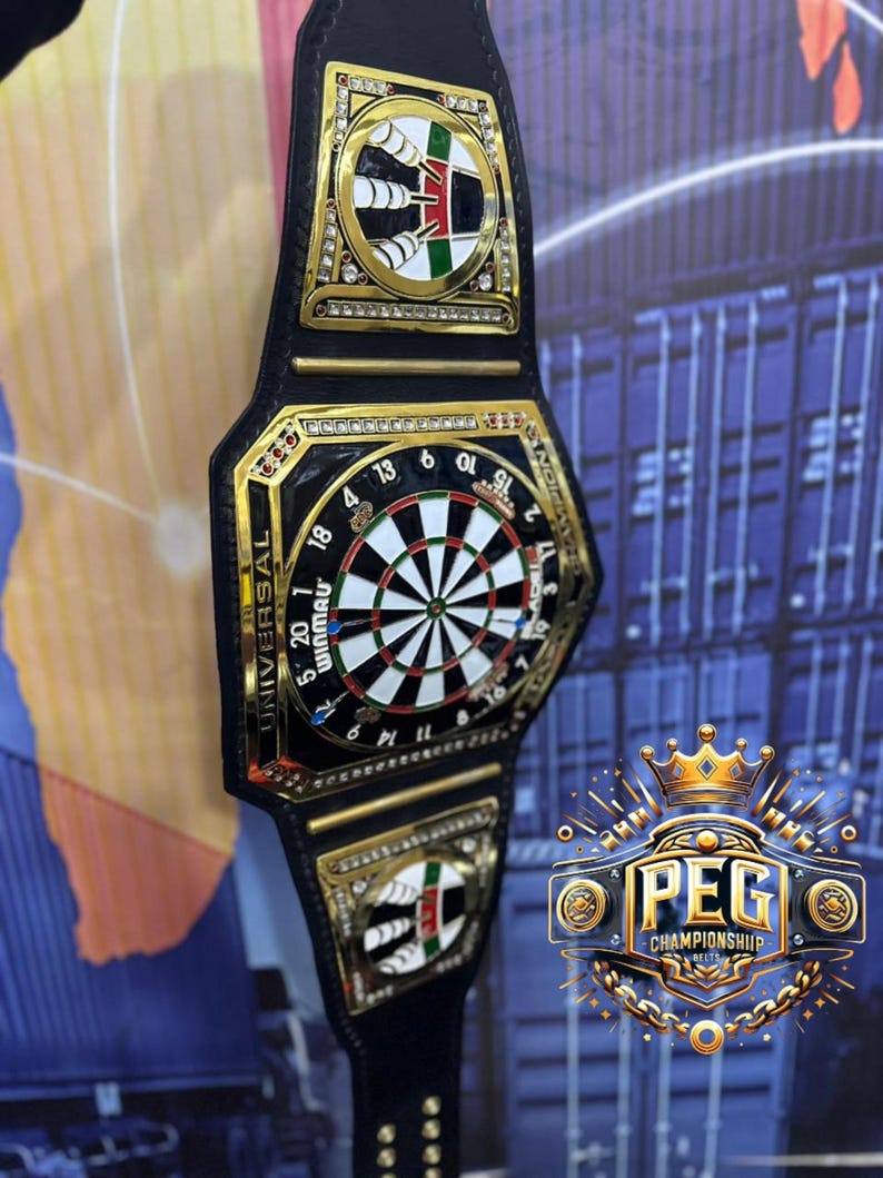May include: A black and gold championship belt featuring a dartboard design at the center. The belt has gold accents, decorative emblems, and the text "UNIVERSAL WINMAU". The image also includes the text "PEG CHAMPIONSHIP BELTS".