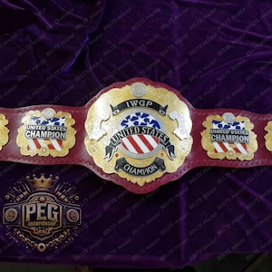 Może przedstawiać: Czerwono-złoty pas mistrzowski wrestlingu z napisem "IWGP United States Champion" na centralnej płytce. Pas ma trzy boczne płytki z napisem "United States Champion" i wzorem globusa.