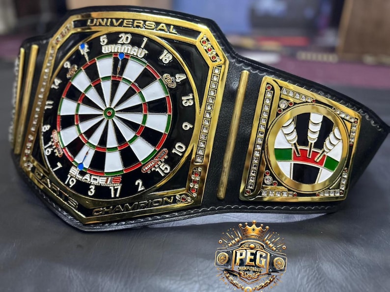 May include: A black and gold championship belt featuring a dartboard design in the center. The belt has the text "UNIVERSAL" and "CHAMPION" on it. The belt also has a logo that says "PEG CHAMPIONSHIP".