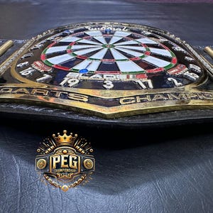 May include: A championship belt with a dartboard design. The belt features a black leather strap with gold-colored accents and a central emblem that reads "DARTS CHAMPION". The dartboard is in the center, with the numbers and colors clearly visible.