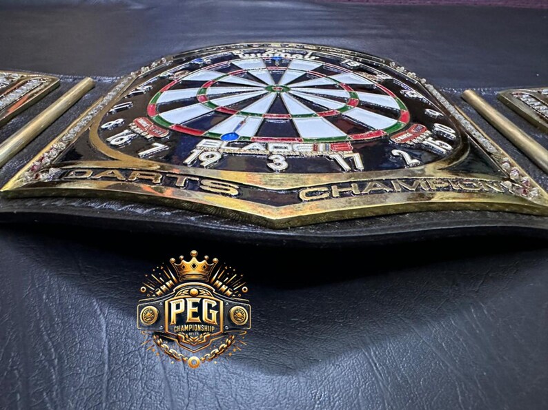 May include: A championship belt with a dartboard design. The belt features a black leather strap with gold-colored accents and a central emblem that reads "DARTS CHAMPION". The dartboard is in the center, with the numbers and colors clearly visible.