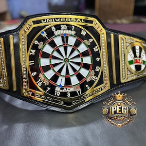May include: A black and gold darts champion belt with a dartboard design at the center. The belt features the text "UNIVERSAL DARTS CHAMPION" and the "PEG CHAMPIONSHIPS" logo. The belt is decorated with rhinestones.