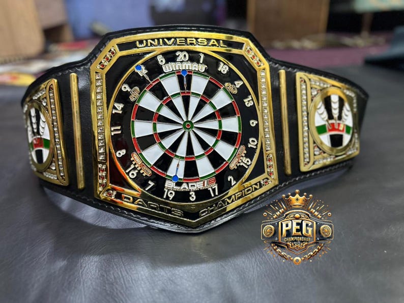 May include: A black and gold darts champion belt with a dartboard design at the center. The belt features the text "UNIVERSAL DARTS CHAMPION" and the "PEG CHAMPIONSHIPS" logo. The belt is decorated with rhinestones.