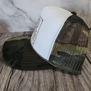 Pode incluir: Bon&eacute; de caminhoneiro com painel frontal branco e um patch retangular marrom e dourado com texto. A aba &eacute; camuflada e a parte de tr&aacute;s em malha &eacute; marrom. O chap&eacute;u tem um bot&atilde;o preto na parte superior e uma borda verde.