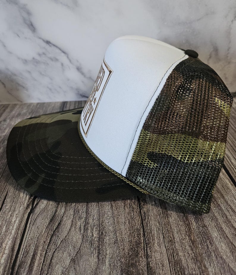 Pode incluir: Bon&eacute; de caminhoneiro com painel frontal branco e um patch retangular marrom e dourado com texto. A aba &eacute; camuflada e a parte de tr&aacute;s em malha &eacute; marrom. O chap&eacute;u tem um bot&atilde;o preto na parte superior e uma borda verde.