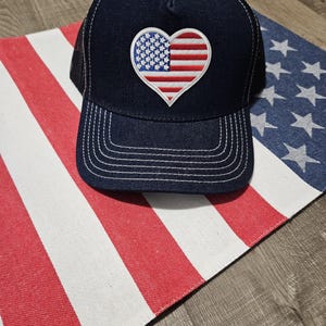 Pode incluir: Um boné de caminhoneiro azul marinho com uma parte de trás de malha e um remendo em forma de coração com a bandeira americana.