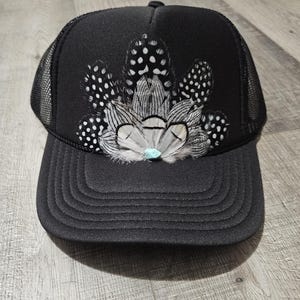 K&ouml;nnte beinhalten: Schwarze Trucker-Kappe mit schwarzem Netzr&uuml;cken. Die Kappe ist mit einem B&uuml;schel wei&szlig;er und schwarzer Federn verziert, in deren Mitte ein t&uuml;rkisfarbener Stein sitzt.