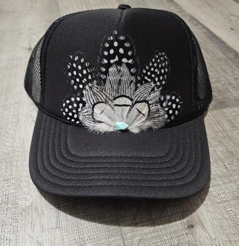 K&ouml;nnte beinhalten: Schwarze Trucker-Kappe mit schwarzem Netzr&uuml;cken. Die Kappe ist mit einem B&uuml;schel wei&szlig;er und schwarzer Federn verziert, in deren Mitte ein t&uuml;rkisfarbener Stein sitzt.