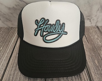 Op de afbeelding: Zwarte en witte truckerpet met het woord "Handy" in turquoise script op de voorkant. De pet heeft een zwarte klep en mesh achterkant, en een wit voorpaneel.