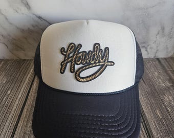 Op de afbeelding: Een marineblauwe en witte truckerpet met het woord "Howdy" in een gouden lettertype in touwstijl. De pet heeft een wit voorpaneel, een marineblauwe rand en een mesh achterkant. De pet ligt op een houten oppervlak.