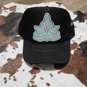 Puede incluir: Gorra de camionero negra con un parche decorativo. El parche presenta un diseño blanco, turquesa y azul con detalles de pedrería. La gorra tiene una parte trasera de malla y una visera curva.