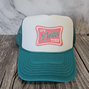 Op de afbeelding: Een turquoise en witte truckerpet met een gebogen klep. De pet heeft een wit voorpaneel met een roze en rode rechthoekige patch met de tekst "It's Lake Time". De pet heeft een turquoise mesh achterkant en een turquoise klep.