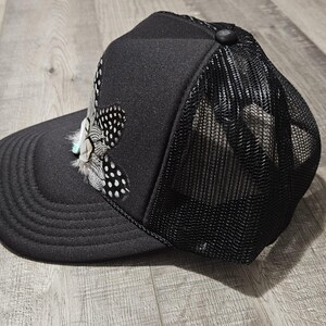 K&ouml;nnte beinhalten: Eine schwarze Trucker-Kappe mit Netzr&uuml;cken und einem gefiederten Design auf der Vorderseite.