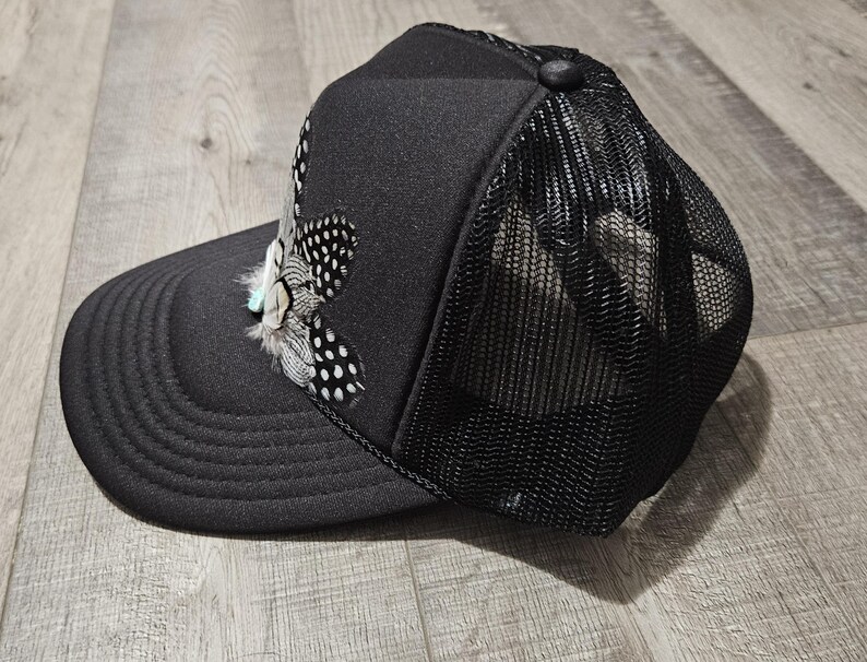 K&ouml;nnte beinhalten: Eine schwarze Trucker-Kappe mit Netzr&uuml;cken und einem gefiederten Design auf der Vorderseite.