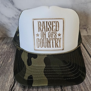 Pode incluir: Bon&eacute; de caminhoneiro com painel frontal branco e estampa camuflada na aba e na parte inferior. O bon&eacute; apresenta o texto bordado "RAISED ON 90'S COUNTRY" em letras douradas dentro de uma borda dourada. O bon&eacute; tem uma parte traseira de malha e um detalhe de corda.