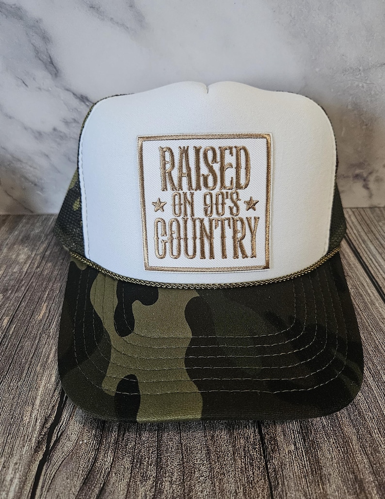 Pode incluir: Bon&eacute; de caminhoneiro com painel frontal branco e estampa camuflada na aba e na parte inferior. O bon&eacute; apresenta o texto bordado "RAISED ON 90'S COUNTRY" em letras douradas dentro de uma borda dourada. O bon&eacute; tem uma parte traseira de malha e um detalhe de corda.