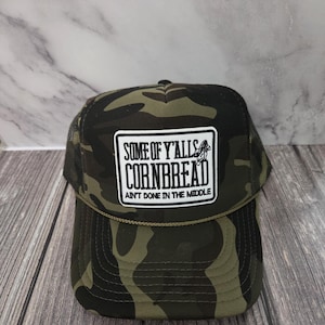 May include: Camouflage trucker hat with a white rectangular patch featuring the text "SOME OF Y'ALLS CORNBREAD AIN'T DONE IN THE MIDDLE". The hat has a mesh back and a curved brim.