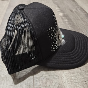 K&ouml;nnte beinhalten: Eine schwarze Mesh-Trucker-Kappe mit einem schwarz-wei&szlig;en Muster und einem t&uuml;rkisfarbenen Feder-Akzent.